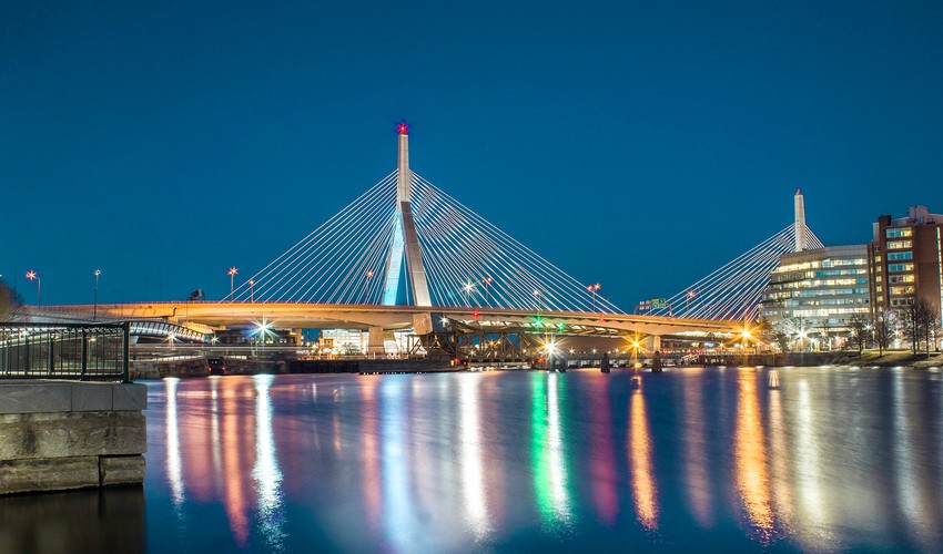 Night view of Boston’s Zakim Bridge with colorful lights reflecting on the Charles River.